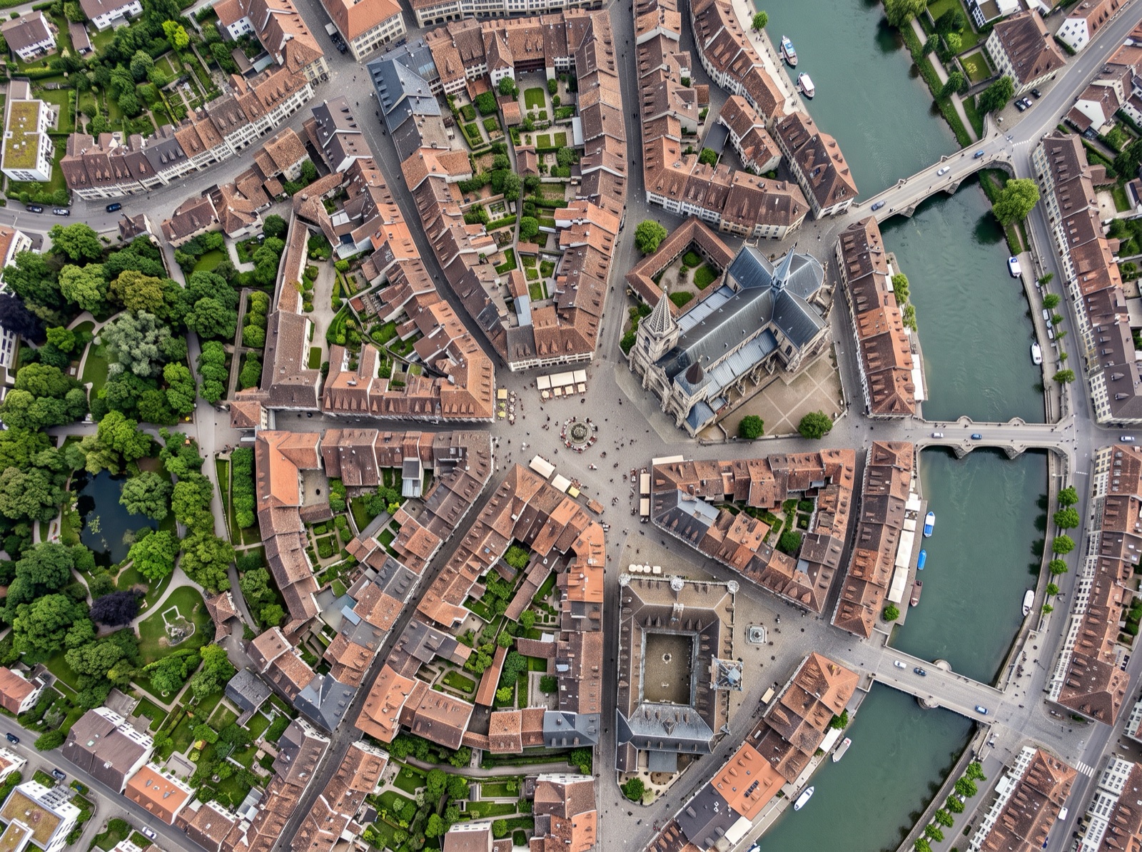 Top-down satellite view of a historic European town centre today — winding streets, tight blocks of tiled roofs, a cathedral, a river with stone bridges and green town gardens